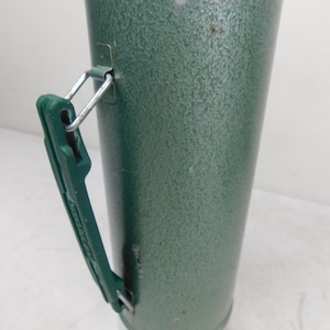 May include: A vintage green metal thermos with a textured hammer-tone finish. The cylindrical container features a dark green plastic side handle attached with silver metal hardware and a silver-colored metal base at the bottom of the unit.