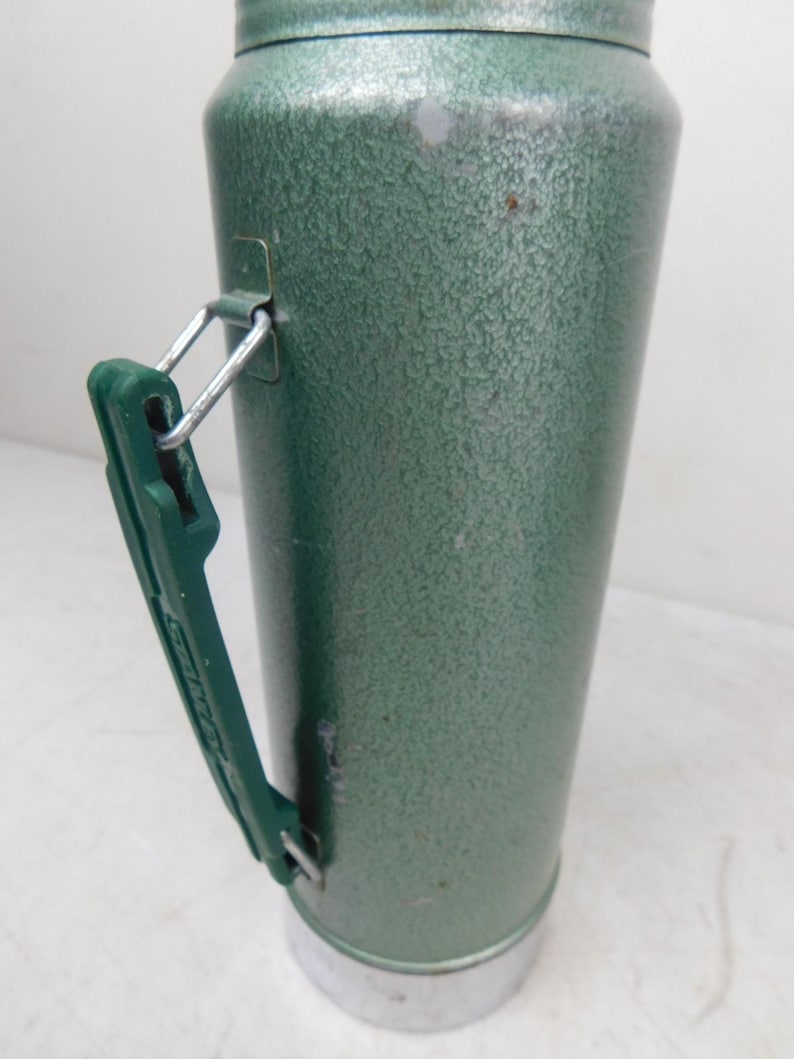 May include: A vintage green metal thermos with a textured hammer-tone finish. The cylindrical container features a dark green plastic side handle attached with silver metal hardware and a silver-colored metal base at the bottom of the unit.
