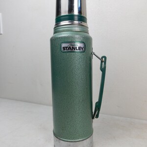 May include: A vintage green Stanley Aladdin thermos with a textured finish, a silver metal cap, and a silver metal base. The body features a green plastic side handle and a rectangular label with the text Aladdin STANLEY in black and white.