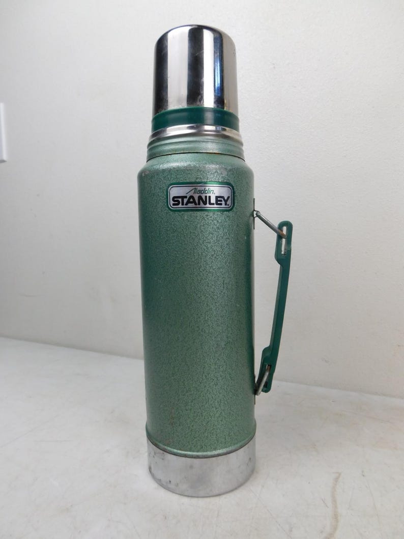 May include: A vintage green Stanley Aladdin thermos with a textured finish, a silver metal cap, and a silver metal base. The body features a green plastic side handle and a rectangular label with the text Aladdin STANLEY in black and white.