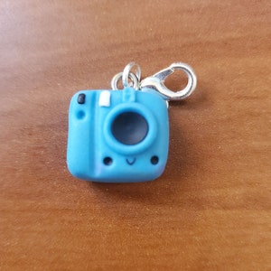 May include: A blue camera charm with a happy face, a white flash, and a black button. The charm has a silver clip.