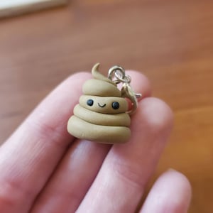 May include: A small brown poop emoji charm with a smiling face. The charm is attached to a silver metal loop.