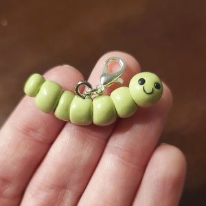 Könnte beinhalten: Ein grüner Wurm-Anhänger mit einem lächelnden Gesicht, aus Polymer Clay-Perlen gefertigt. Der Anhänger hat einen silbernen Metallverschluss.