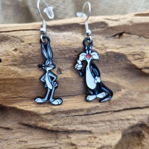 Könnte beinhalten: Ohrringe zum Aufhängen mit den Zeichentrickfiguren Bugs Bunny und Sylvester the Cat. Der Bugs Bunny-Ohrring ist grau und weiß, während der Sylvester-Ohrring schwarz und weiß mit einer roten Nase ist. Beide Ohrringe haben silberfarbene Haken.