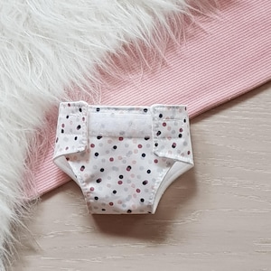 Könnte beinhalten: Eine weiße Windel mit einem Tupfenmuster in den Farben Rosa, Grau und Schwarz. Die Windel hat einen weißen Klettverschluss.