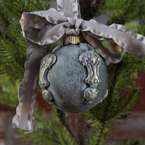 May include: A gray Christmas ornament with gold accents and a gray satin ribbon. The ornament is shaped like a sphere and has a textured surface.