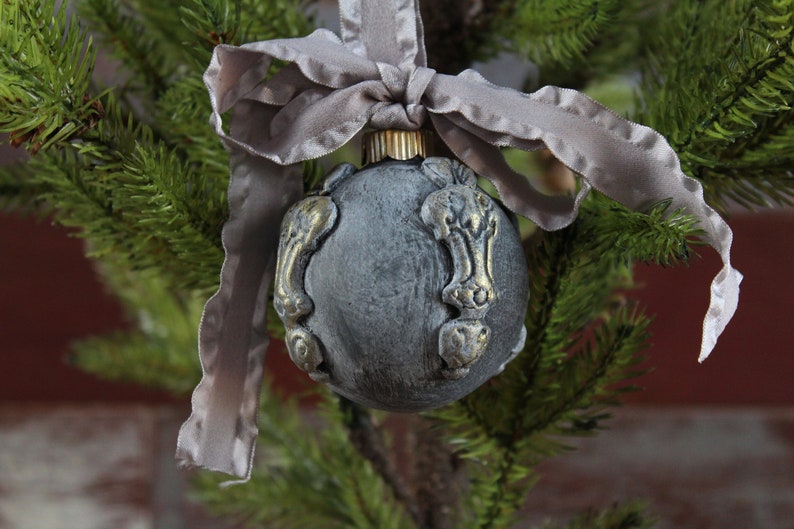 May include: A gray Christmas ornament with gold accents and a gray satin ribbon. The ornament is shaped like a sphere and has a textured surface.