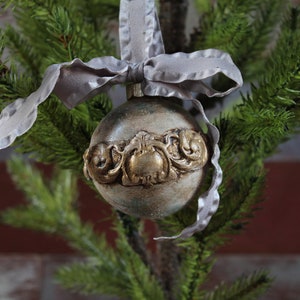 May include: A silver and gold Christmas ornament with a decorative design. The ornament is hanging from a gray ribbon on a green artificial Christmas tree.