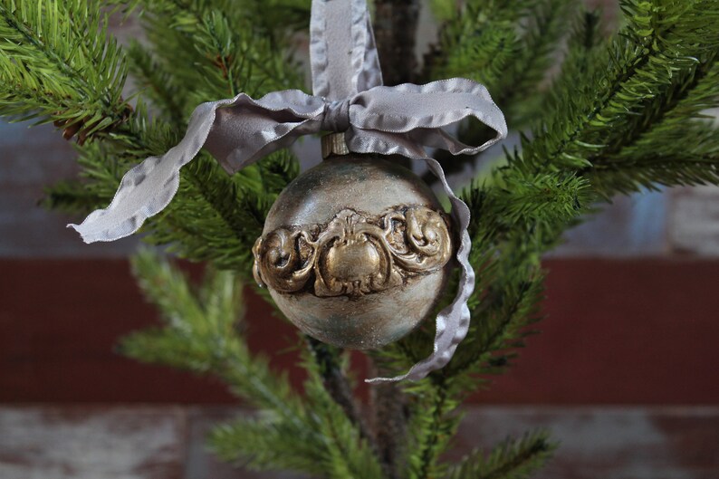 May include: A silver and gold Christmas ornament with a decorative design. The ornament is hanging from a gray ribbon on a green artificial Christmas tree.