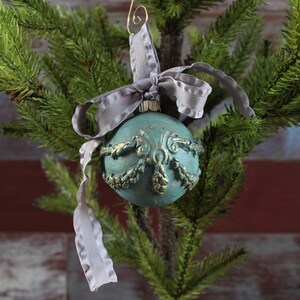 May include: A teal blue Christmas ornament with gold accents and a gray ribbon. The ornament is hanging on a green artificial Christmas tree.