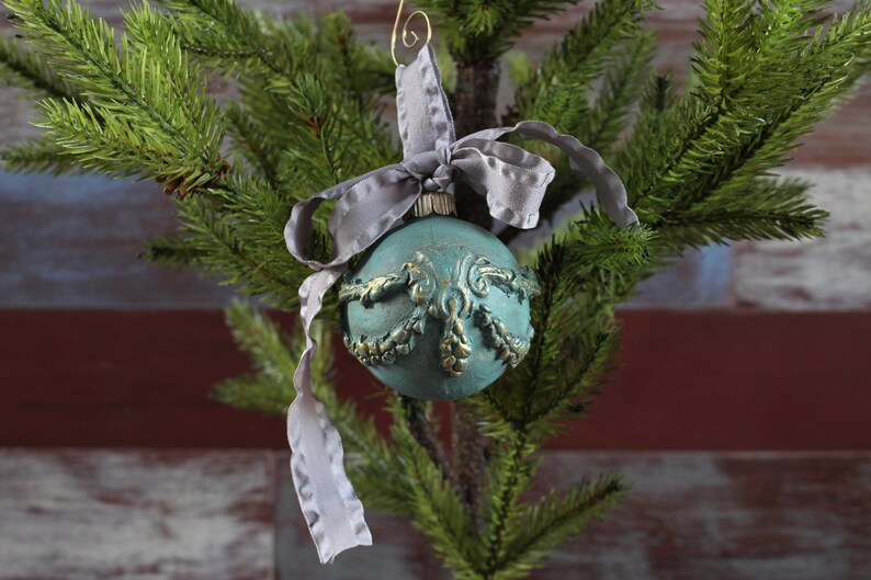 May include: A teal blue Christmas ornament with gold accents and a gray ribbon. The ornament is hanging on a green artificial Christmas tree.