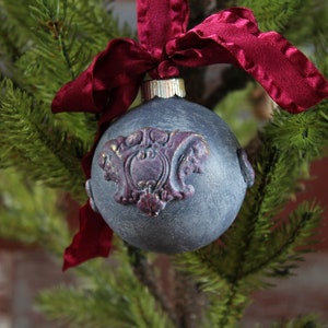 May include: A gray Christmas ornament with a burgundy and gold design, decorated with a burgundy velvet ribbon.