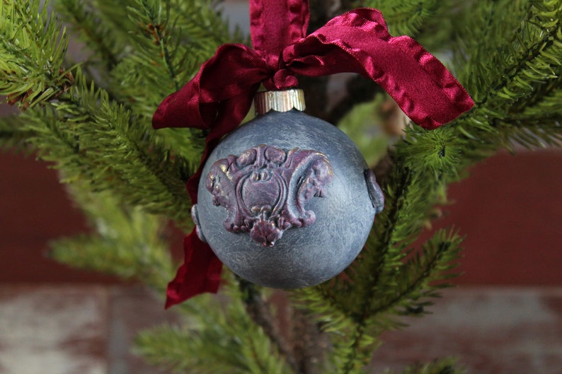 May include: A gray Christmas ornament with a burgundy and gold design, decorated with a burgundy velvet ribbon.