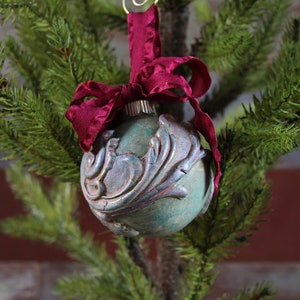 May include: A teal blue glass Christmas ornament with a silver and gold floral design, accented with a burgundy velvet ribbon.