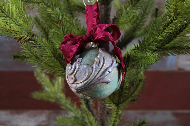 May include: A teal blue glass Christmas ornament with a silver and gold floral design, accented with a burgundy velvet ribbon.
