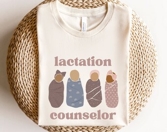 May include: A beige t-shirt with the text "lactation counselor" in pink lettering. Four illustrations of babies wrapped in blankets are above the text.