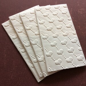 Könnte beinhalten: Ein Stapel cremefarbener Papierservietten mit einem geprägten Herzmuster. Jede Serviette zeigt Reihen von Herzen, die durch gepunktete Linien verbunden sind. Die Servietten liegen auf einer dunkelbraunen Oberfläche, die das Design hervorhebt.