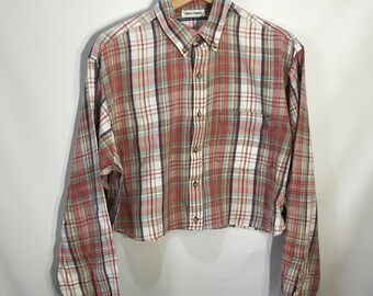 Puede incluir: Una camisa de botones a cuadros recortada con mangas largas. La camisa es de cuadros rojos, blancos, azules y verdes. La camisa tiene un cierre de botones en la parte delantera y un solo bolsillo en el pecho izquierdo. La etiqueta de la camisa dice "Urban Threads".