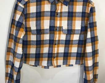 Puede incluir: Una camisa de franela de manga larga y corte corto con un estampado de cuadros en amarillo, azul y blanco. La camisa tiene un cierre de botones en la parte delantera y dos bolsillos en el pecho. La etiqueta de la camisa dice "Urban Thread".
