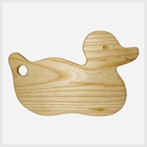 Könnte beinhalten: Ein hölzernes Schneidebrett in Form einer Ente. Das hellfarbene Holz zeigt natürliche Maserungen. Es hat ein Loch in der Nähe des Schwanzes und ein lächelndes Gesicht. Das Brett ist etwa 30 cm lang.