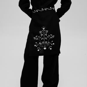 Op de afbeelding: Een zwarte set bestaande uit een shirt met lange mouwen en een wijde broek, voorzien van een wit geborduurd bloemmotief op het voorpand. Het ontwerp bevat een horizontale golvende lijn bij de taille en een symmetrisch botanisch patroon.
