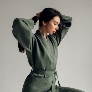 May include: A woman wearing a sage green linen jumpsuit with a self-tie belt. The jumpsuit has a relaxed fit and a v-neckline.