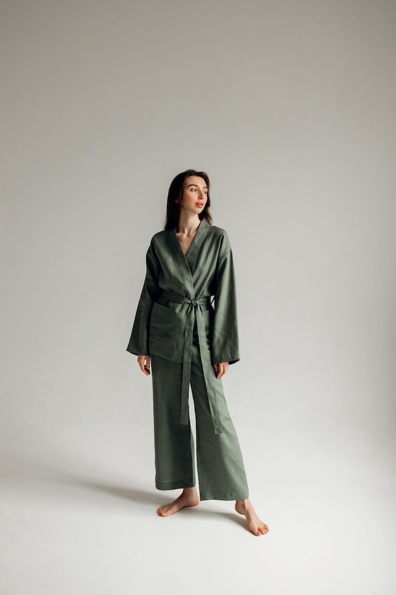 May include: A woman wearing a green linen pantsuit with a long-sleeved jacket and wide-leg pants. The jacket has a tie belt and a v-neck.