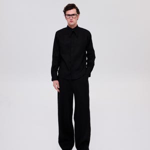May include: A man wearing a black long-sleeved shirt with a collar and black pants. He is wearing black-framed glasses.