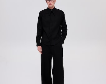 Op de afbeelding: Een zwart overhemd met lange mouwen en een puntige kraag, gecombineerd met een bijpassende zwarte broek met wijde pijpen. De outfit wordt gedragen met een zwarte bril met dik montuur, wat zorgt voor een monochrome look met strakke lijnen.