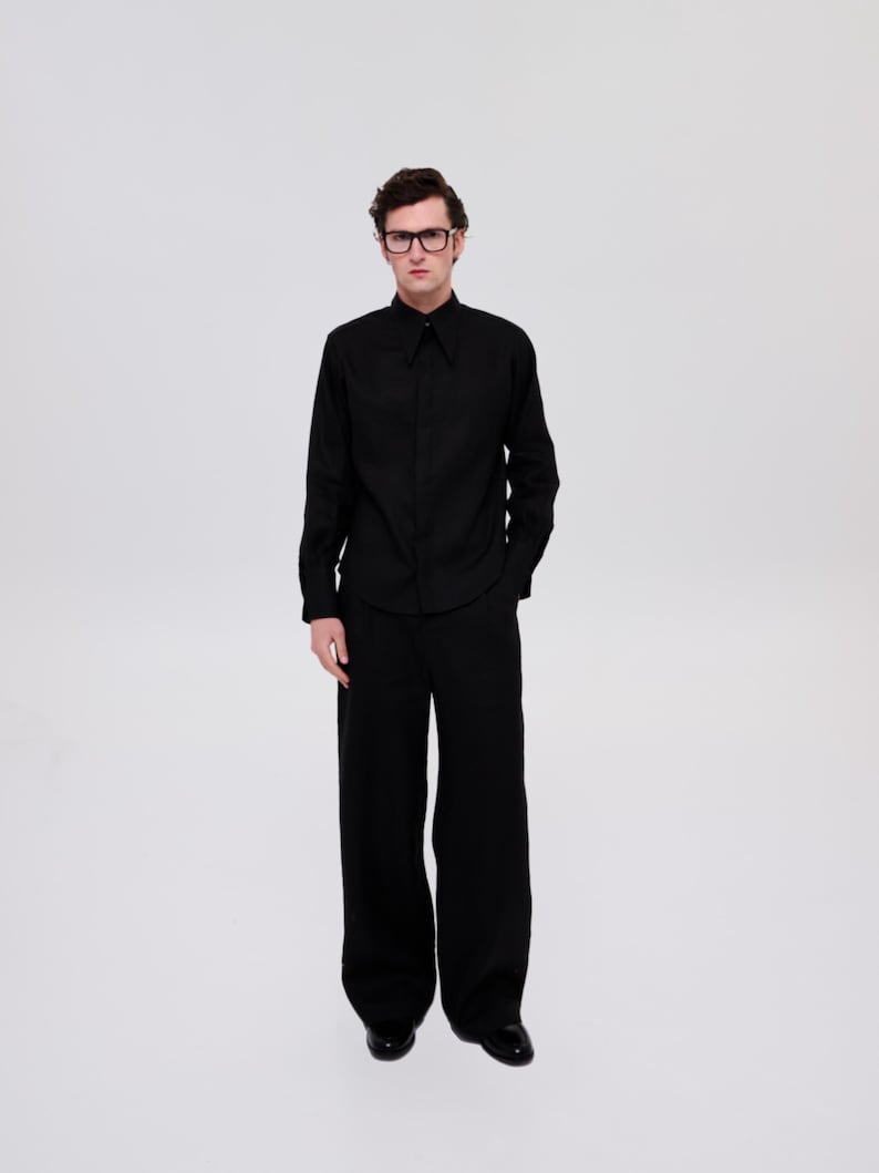 May include: A man wearing a black long-sleeved shirt with a collar and black pants. He is wearing black-framed glasses.