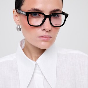May include: Black-framed eyeglasses with a thick, rectangular silhouette and clear lenses. The temples feature a silver-toned metallic detail near the hinge. The right lens is etched with the text SAINT LAURENT PARIS in a small, clean font.