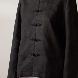 May include: Black linen jacket with a mandarin collar and long sleeves. The jacket features decorative frog closures down the front. The fabric has a subtle texture, and the overall design is simple and elegant.
