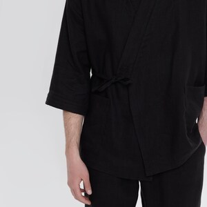 May include: A black linen kimono with a long sleeve and a tie belt. The kimono is worn over black pants.