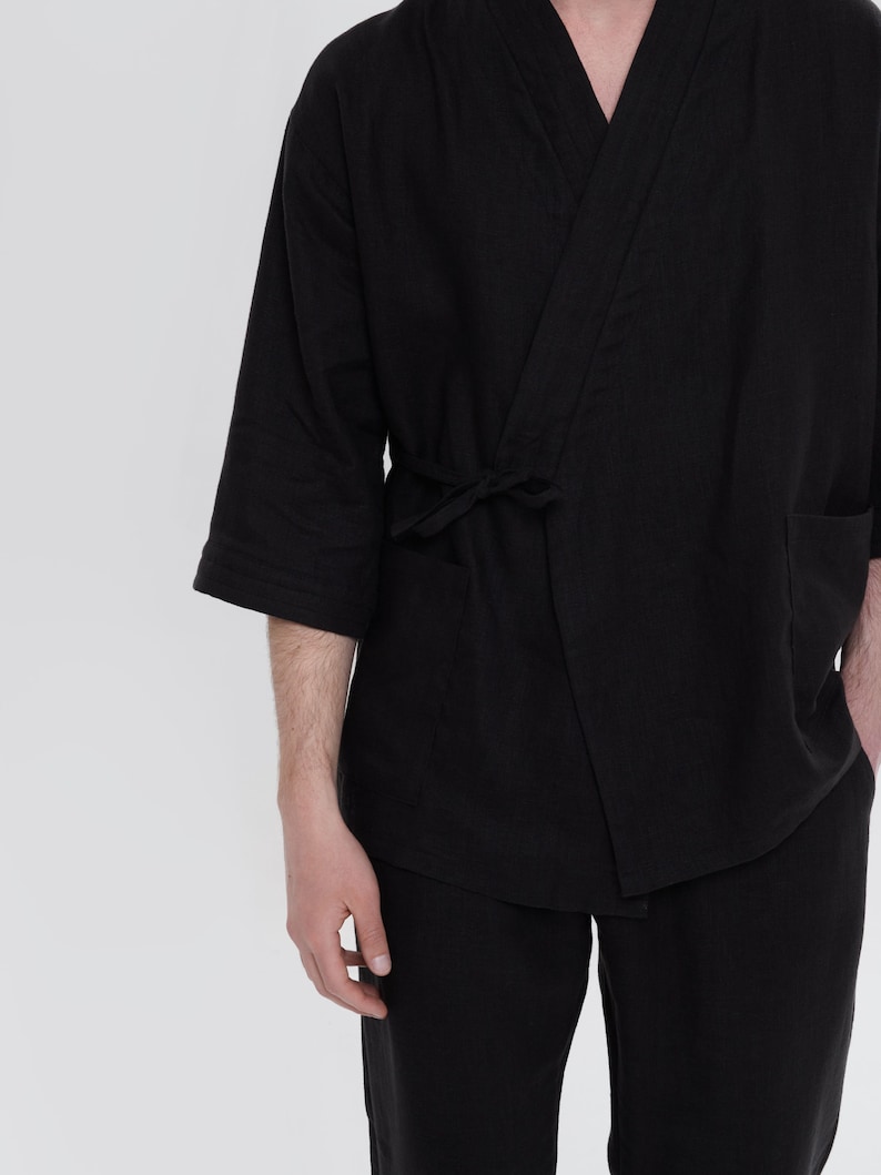 May include: A black linen kimono with a long sleeve and a tie belt. The kimono is worn over black pants.