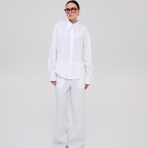 May include: A crisp white long-sleeved button-down shirt with a pointed collar and wide cuffs, paired with matching white wide-leg trousers featuring large front pockets. The ensemble is completed with black pointed-toe shoes and black-rimmed glasses.