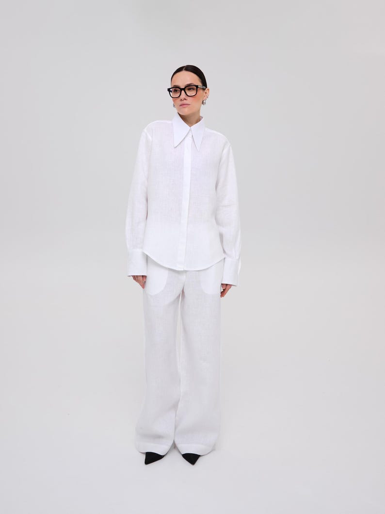 May include: A crisp white long-sleeved button-down shirt with a pointed collar and wide cuffs, paired with matching white wide-leg trousers featuring large front pockets. The ensemble is completed with black pointed-toe shoes and black-rimmed glasses.
