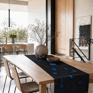 May include: A wooden dining table with a black table runner featuring a blue celestial design. The table is set with wooden bowls and a large vase with dried branches. The chairs have wooden seats and black metal legs.