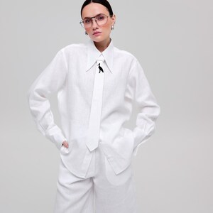 May include: A white long-sleeved button-down shirt with a pointed collar and a matching white necktie featuring a small black bird graphic. The shirt is paired with matching white knee-length shorts, creating a coordinated monochromatic outfit.