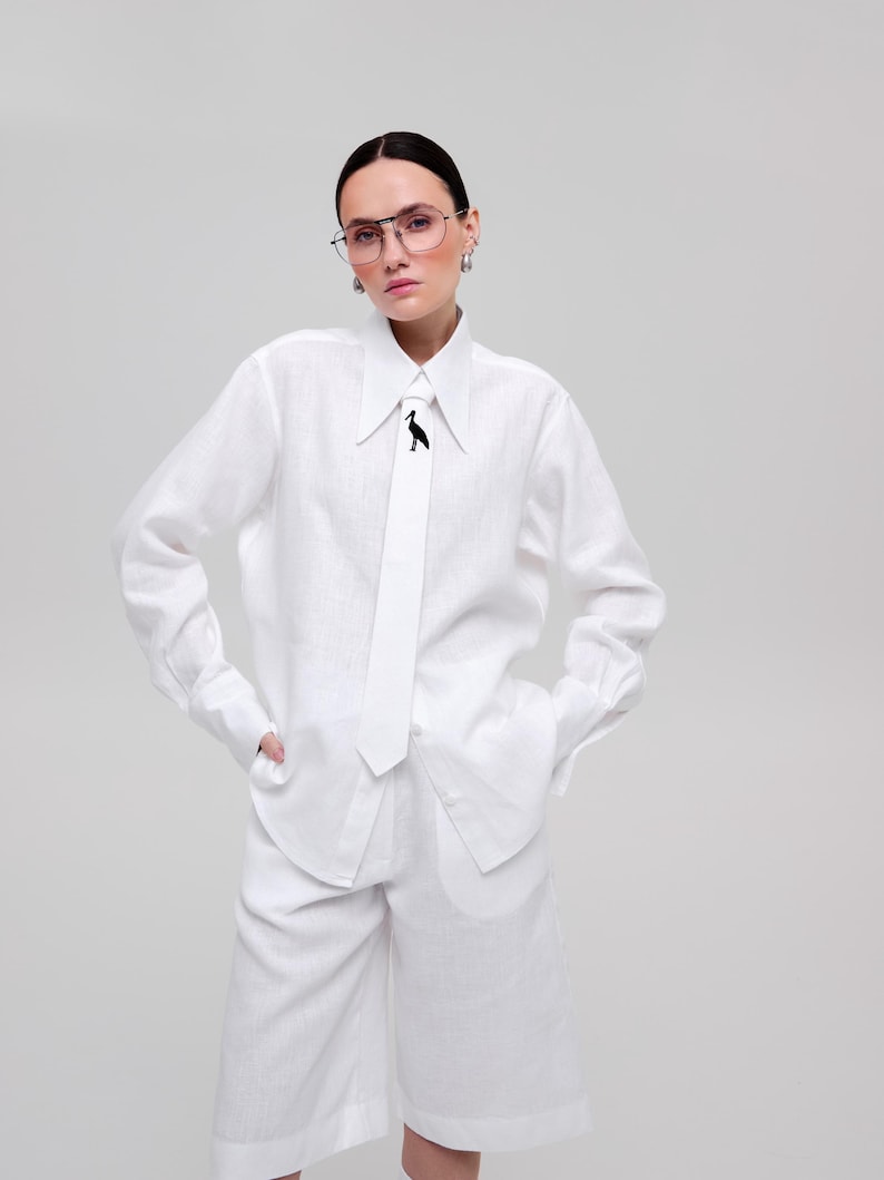 May include: A white long-sleeved button-down shirt with a pointed collar and a matching white necktie featuring a small black bird graphic. The shirt is paired with matching white knee-length shorts, creating a coordinated monochromatic outfit.