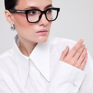 May include: Black rectangular eyeglasses with thick frames and clear lenses. The frames feature a silver-toned metal detail on the temple hinge. The right lens has the Saint Laurent Paris logo printed in small white text on the upper corner.