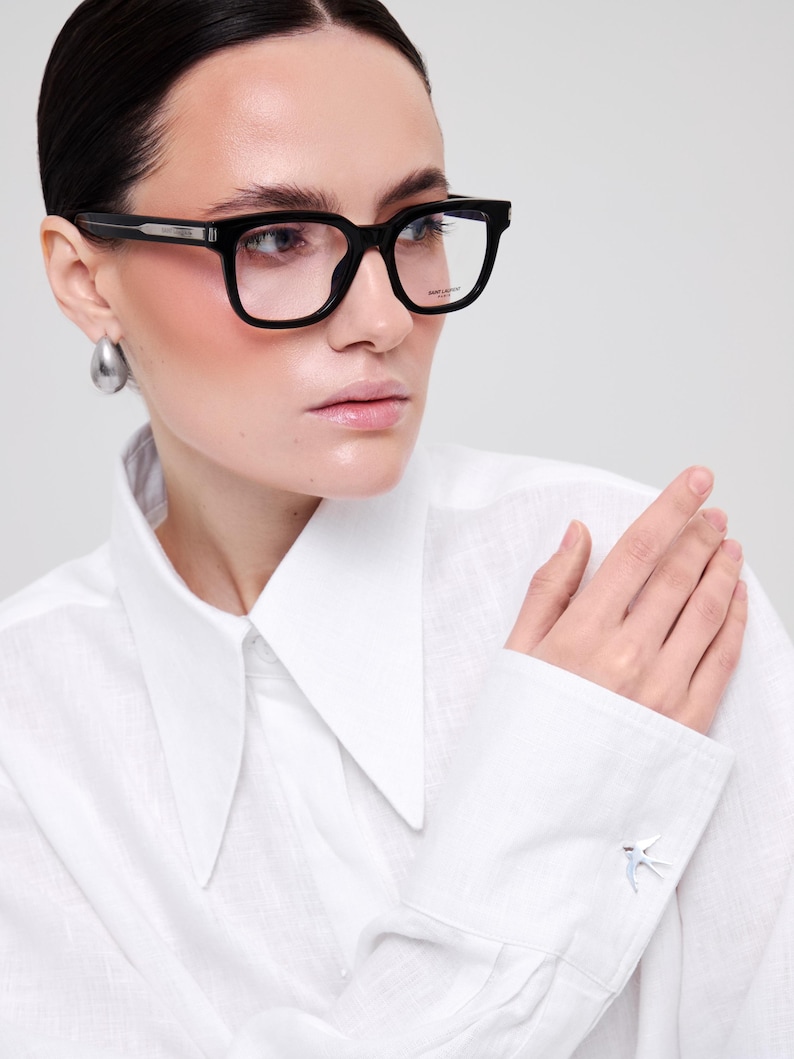 May include: Black rectangular eyeglasses with thick frames and clear lenses. The frames feature a silver-toned metal detail on the temple hinge. The right lens has the Saint Laurent Paris logo printed in small white text on the upper corner.