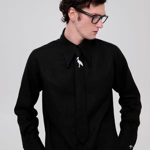 May include: A black long-sleeved shirt with a white bird design on the collar. The shirt has a long, thin tie with a pointed end. The person wearing the shirt is also wearing black-framed glasses.