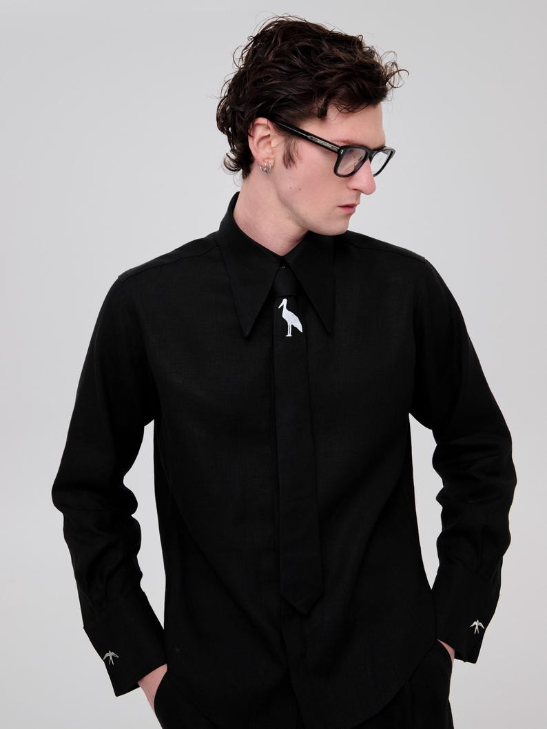 May include: A black long-sleeved shirt with a white bird design on the collar. The shirt has a long, thin tie with a pointed end. The person wearing the shirt is also wearing black-framed glasses.