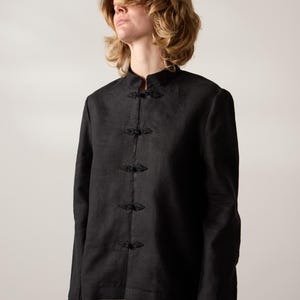 May include: A black linen jacket featuring a mandarin collar and frog fastenings. The long-sleeved jacket is buttoned up and worn over white trousers. The jacket has a simple, elegant design.