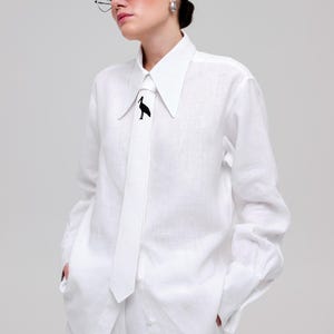 May include: A white linen shirt with a long sleeve and a white tie featuring a black bird design. The shirt has a pointed collar and a relaxed fit.