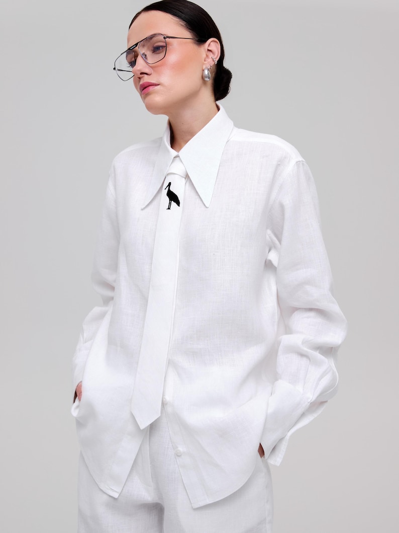 May include: A crisp white long-sleeved button-down shirt with a pointed collar and a matching white necktie. The necktie features a small, black, minimalist graphic of a standing stork bird positioned centrally near the top of the tie.