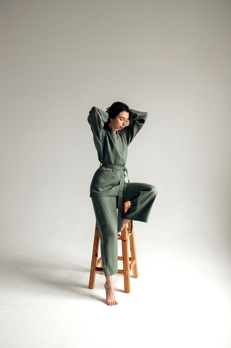 May include: A woman sits on a wooden stool wearing a sage green linen jumpsuit with a matching belt.