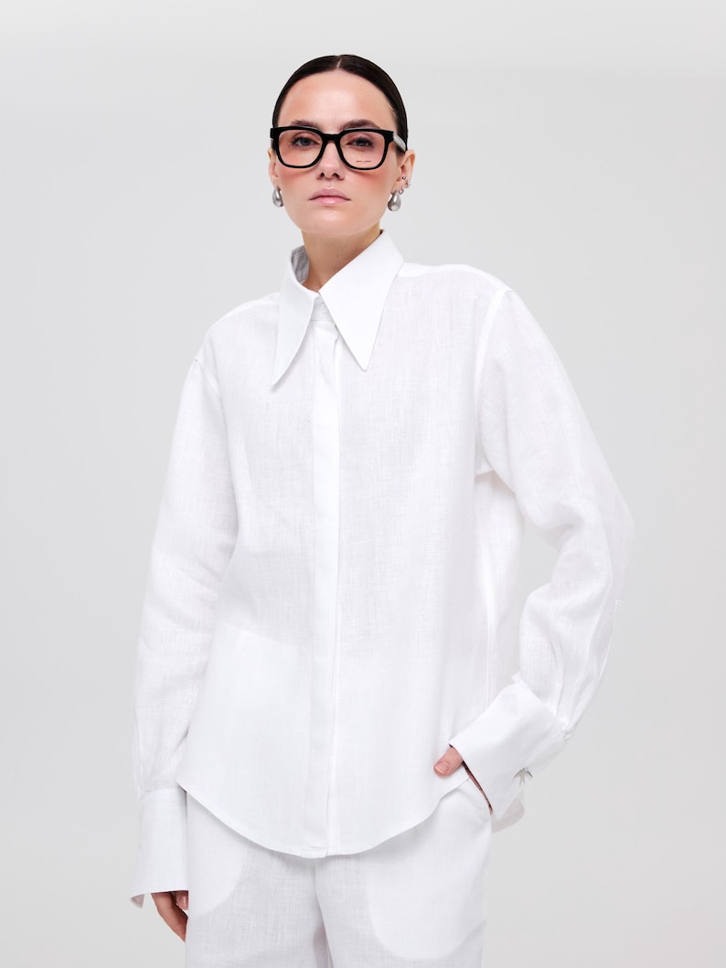 May include: A crisp white long-sleeved button-down shirt with a pointed collar and wide cuffs. The shirt is paired with matching white trousers. The look is completed with thick-rimmed black glasses and silver drop earrings.