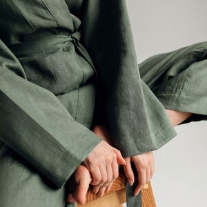 May include: A woman wearing a green linen jumpsuit with a sash tied at the waist. She is sitting on a wooden stool with her legs crossed.