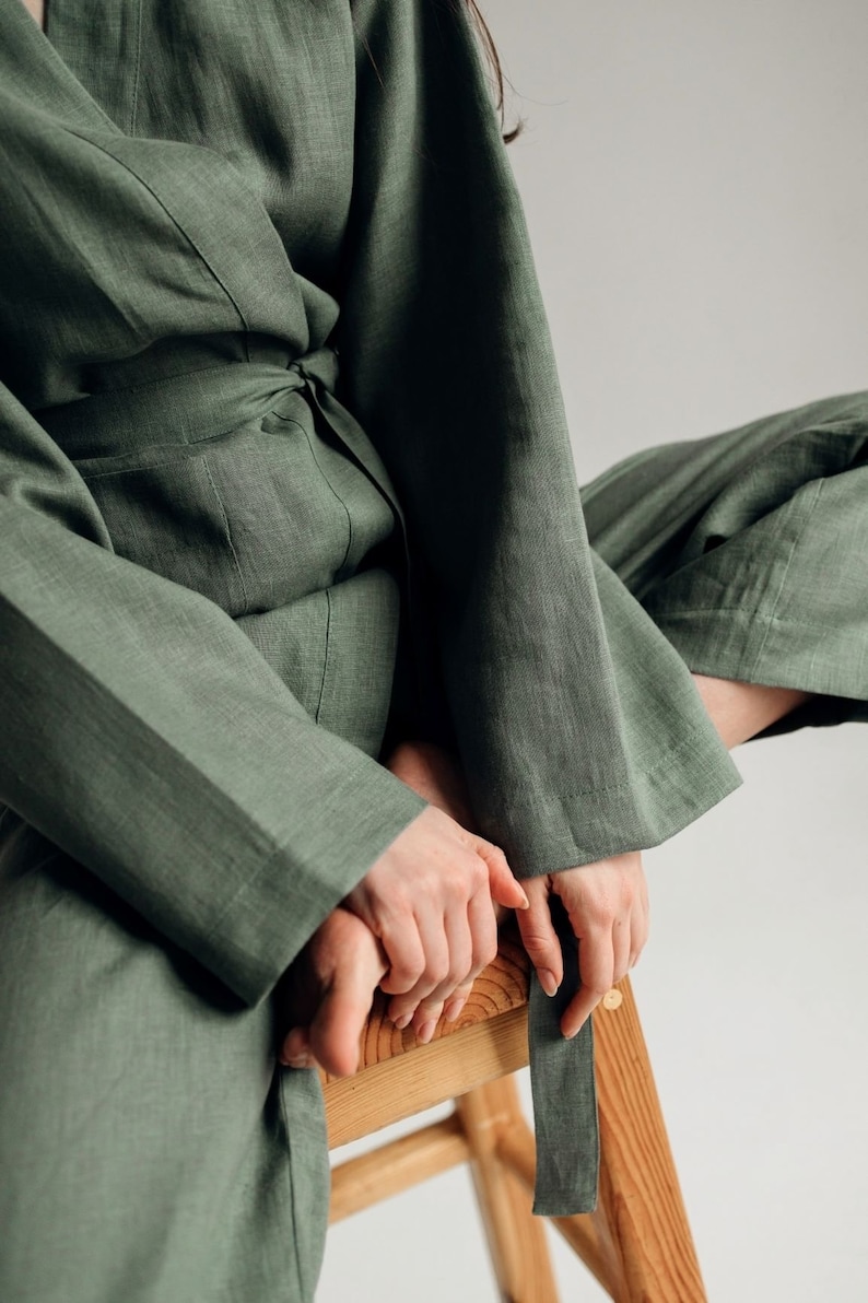 May include: A woman wearing a green linen jumpsuit with a sash tied at the waist. She is sitting on a wooden stool with her legs crossed.